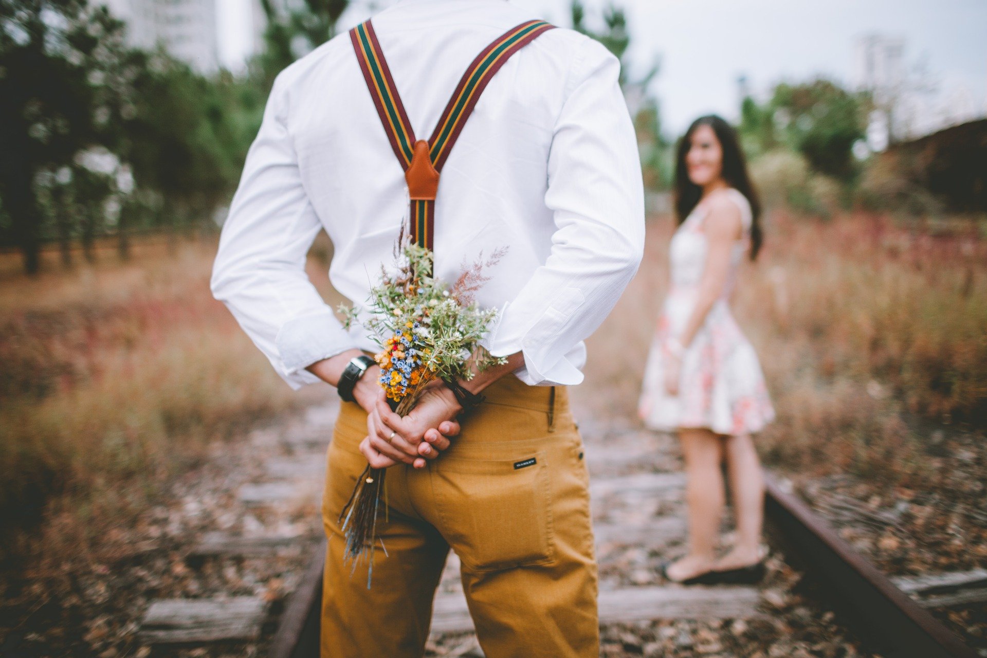 Man holding bouquet of flowers hidden behind his back, woman looking at him ahead.