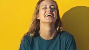 Blond woman in blue blouse smiling on yellow background.